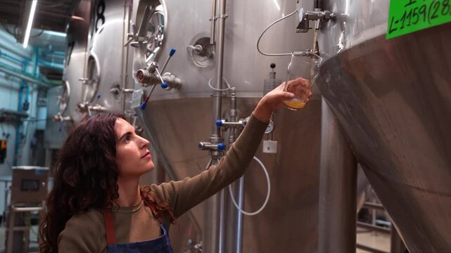 Young woman pouring a fresh glass of beer from a fermentation tank in a brewery. Professional brewmaster checking the quality of her product