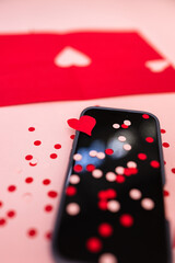 close-up of a cellphone with a black screen with pink and red confetti and a cutout paper heart against a pink background
