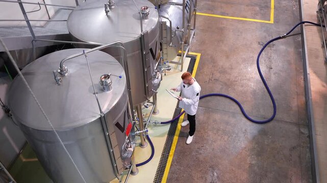 High angle view of a brewmaster inspecting the quality control of beer production, writing on a clipboard next to stainless steel vats