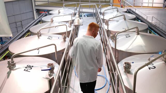 Brewmaster in a white lab coat walking and controlling the brewing process in a modern brewery. High angle view of industrial equipment