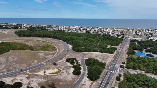 An expansive aerial shot shows a circular road loop around a grassy hill topped by a White Wright Brothers monument, with distant Kill Devil Hills and Outer Banks Coastline under a clear blue sky.