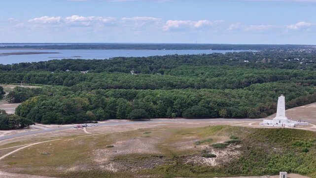 An expansive aerial shot shows a circular road loop around a grassy hill topped by a White Wright Brothers monument, with distant Kill Devil Hills and Outer Banks Coastline under a clear blue sky.