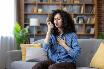 Young woman having difficulty breathing and managing an acute asthma attack at home, using an inhaler to alleviate respiratory symptoms and seeking quick relief