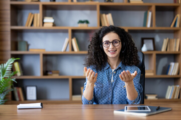 Young woman with headset and glasses smiling and gesturing during a remote video call from her home office, seated at a wooden desk with bookshelf in the background