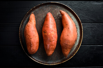 Raw organic sweet potatoes on a plate. Black wooden background.