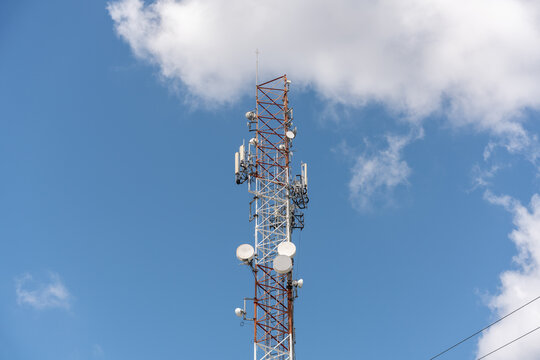 View of a massive telecommunications antenna or transmission tower, supporting various communication antennas for mobile telephony, radio, TV, and internet. The background is a vibrant blue sky. - Powered by Adobe