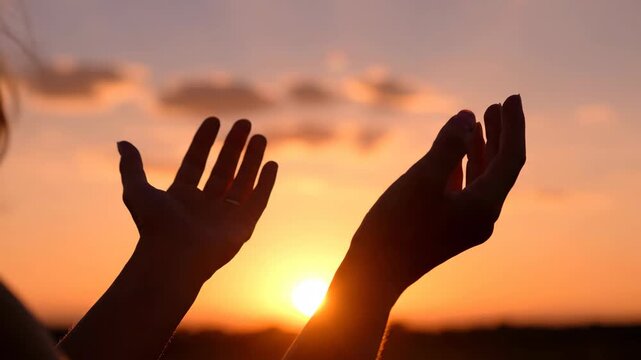 Silhouette of hands raised in prayer against a sunset sky. A person worshiping with faith and hope. Spirituality and gratitude concept