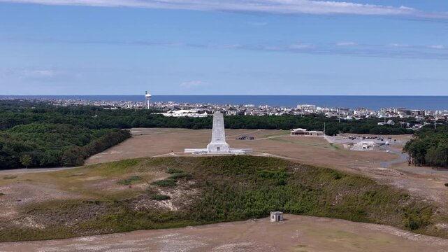 An expansive aerial shot shows a circular road loop around a grassy hill topped by a White Wright Brothers monument, with distant Kill Devil Hills and Outer Banks Coastline under a clear blue sky.