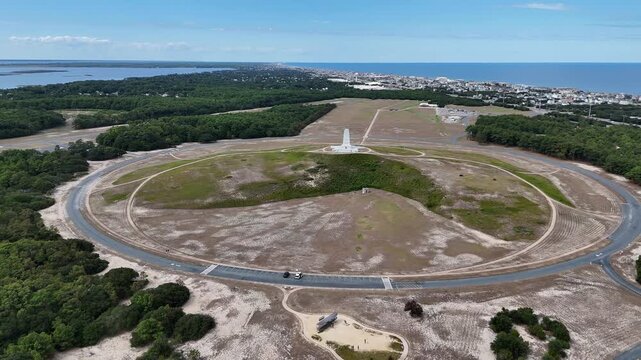 An expansive aerial shot shows a circular road loop around a grassy hill topped by a White Wright Brothers monument, with distant Kill Devil Hills and Outer Banks Coastline under a clear blue sky.