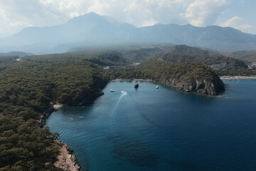Drone photo of a serene mountain bay surrounded by forest with boats on deep blue water under a hazy summer sky. Peaceful coastal landscape. The Gulf of Antalya, Turkey