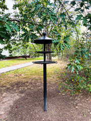 A black metal bird feeder standing in a green park among trees and bushes. Peaceful outdoor scene showing a feeding station for birds in a natural environment.