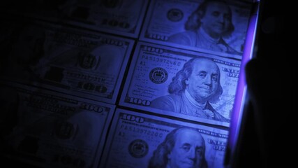 Close up of a handheld ultraviolet detector scanning across neat rows of hundred dollar bills, verifying authenticity by illuminating hidden security features on the banknotes in a dark environment.