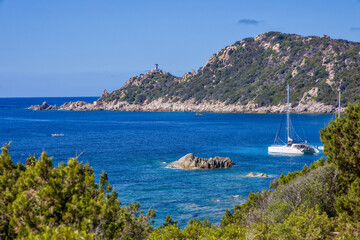 Obraz premium the bay of Cap Muro with its beautiful turquoise water, boats at anchor, and the lighthouse on the hill in sunshine and blue skies