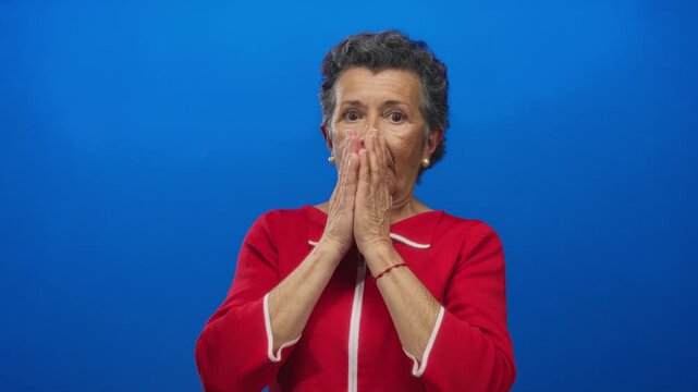Senior woman with grey hair in a red sweater looks shocked against a vibrant blue background, expressing surprise and emotion vividly captured in this stock image.