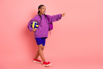 Athletic woman holding a volleyball, dressed in colorful sportswear on a pink background, pointing cheerfully forward.