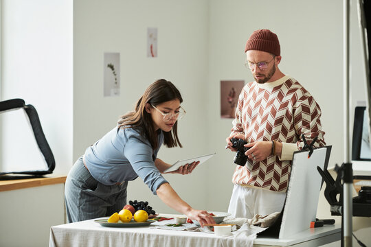 Man holding camera standing beside Asian woman arranging food on table, both collaborating on food photography project in bright studio setting