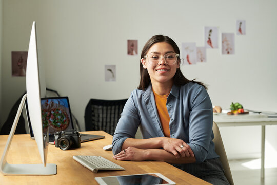 Portrait of woman sitting at desk smiling at camera, using desktop computer with digital tablet and camera on table, creative workspace in background