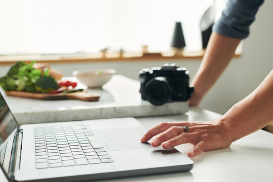 Woman using laptop on kitchen counter with digital camera and fresh vegetables in background, hand resting on trackpad, preparing for food photography