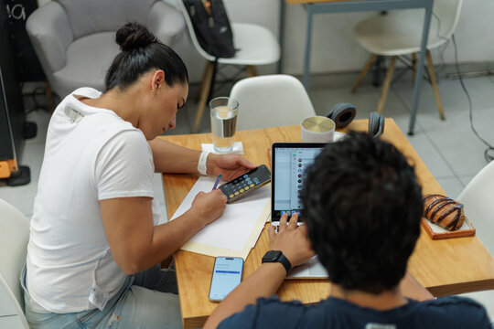 J&oacute;venes profesionales trabajando juntos en una cafeter&iacute;a moderna, mujer calculando en el tel&eacute;fono y hombre usando laptop.
