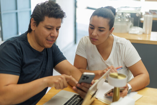 Dos colegas latinos discuten contenido en un smartphone y laptop, colaborando en un espacio de trabajo moderno.