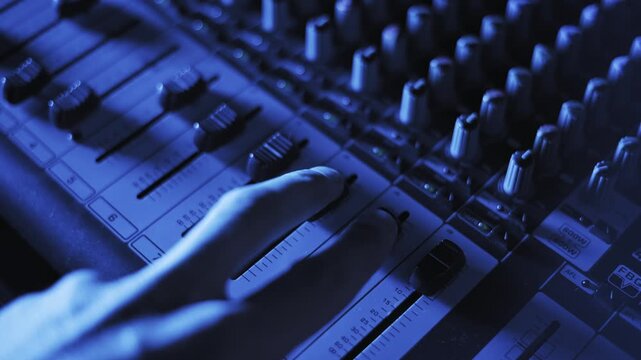 Close angle of a sound technician fingers pushing up a fader on a professional mixing board adjusting channel volume under shifting purple blue and orange stage lights in a dark music studio.