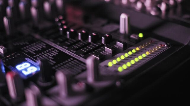 Macro of audio mixer level indicators flashing with green and yellow LEDs faders and a digital display in a professional recording studio with shifting colorful lights for music production.