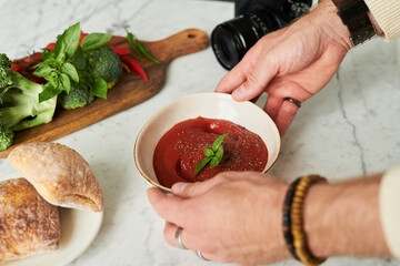 Man holding bowl of tomato soup garnished with basil on marble table, fresh bread and assorted vegetables including broccoli and peppers nearby