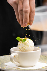 Close-up of a hand sprinkling crushed nuts over vanilla ice cream in a white cup, garnished with mint. Dessert presentation concept with shallow depth of field.