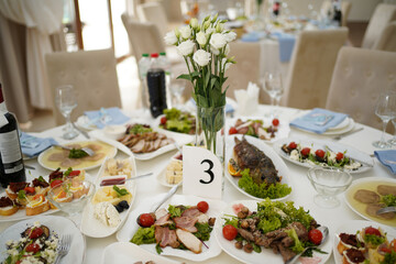 Festive table with various dishes, salads, meat and fish, table setting with flowers in a vase and table number.