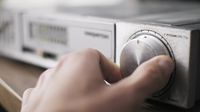 Macro perspective of a hand rotating the master volume dial on an old hi-fi stereo system receiver highlighting the worn brushed aluminum texture and Cyrillic lettering in shallow focus.