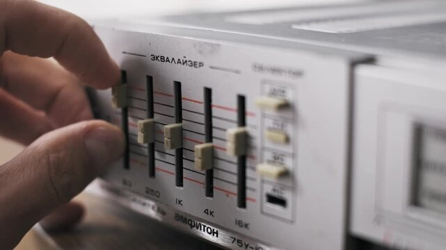 Side angle of fingers adjusting multiple frequency faders on a graphic equalizer of a vintage amplifier tuning sound with beige sliders on a silver metal console with Cyrillic text.