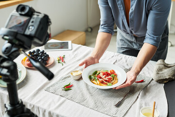 Woman arranging plate of pasta with vegetables on table in front of camera, preparing food for photography or video content creation, hands visible holding dish