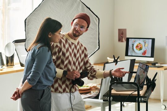 Man wearing glasses showing Asian woman images on laptop during food photography session in studio, both focusing on computer screen together