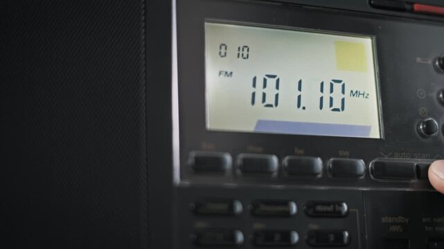 Angled close-up of a finger pressing an auto scan button tuning a digital radio up the FM band. The LCD shows increasing MHz frequency signal strength and timer next to a speaker grill.