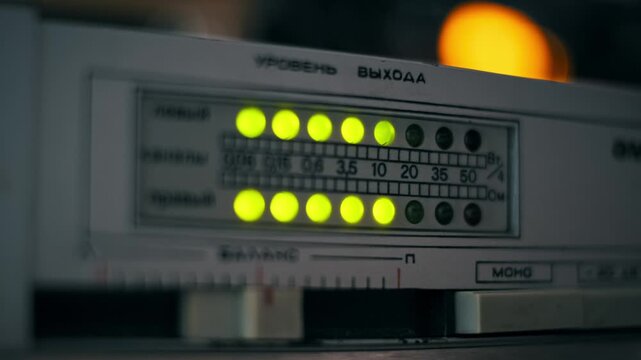 Low-light close-up of a retro audio amplifier's blinking LED level meter showing yellow-green sound indicators with a warm bokeh light in the background labeled with Cyrillic text for Output Level.