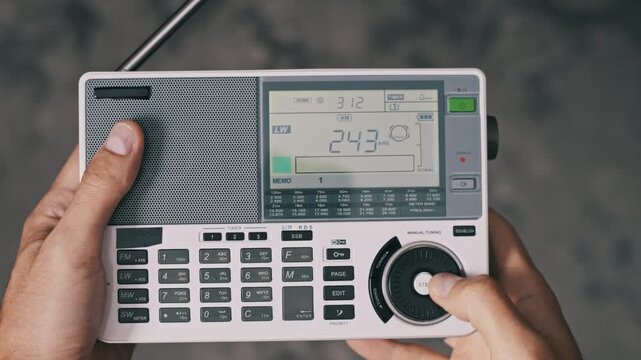 Top-down view of hands operating a multi-band digital radio. The user presses buttons to switch between LW MW SW and FM bands before using the tuning knob to scan for different stations.