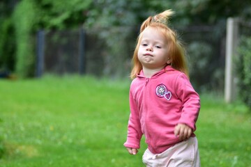 Fototapeta premium Playful red-haired toddler in a pink hoodie running on green grass with a cheerful expression, enjoying outdoor play and fresh air on a bright day in the garden.