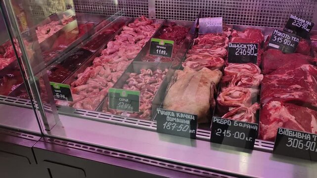 Panning shot of butcher shop refrigerated display case. Various fresh raw meat cuts like veal, lamb ribs, offal, and a whole duck carcass with Cyrillic price tags under cool pink lighting.