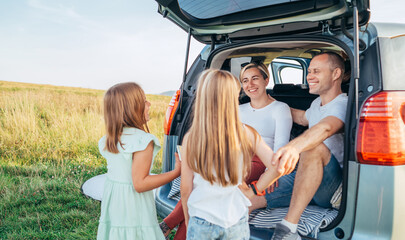 Portrait of Happy young couple with little daughters sitting inside car trunk during auto trop. They are smiling, laughing and chatting. Family values, traveling concept.