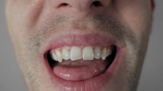 Macro shot of a man with stubble making funny faces, sticking his tongue out, rolling and folding it in unusual ways. He shows his crooked teeth in a goofy, playful, and comical expression.