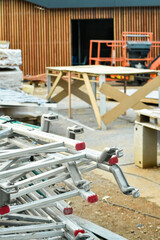 A vertical shot of a construction site with stacked aluminum scaffolding components in the foreground. In the background, wooden walls, sawhorse tables, and other building equipment are visible.