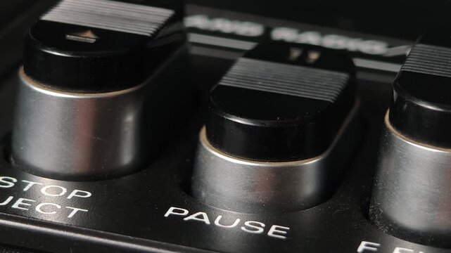 Extreme close up of a finger pressing a pause button on a vintage tape player showing a detailed fingerprint texture black and silver controls and white lettering on a dusty black panel of a retro