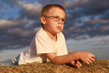A boy with glasses lies on his stomach on a hay bale in golden sunset light under a picturesque sky. The photo is taken from below. Concept for leisure, childhood, copy space background.