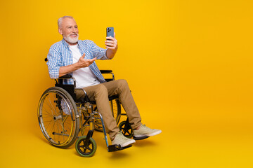 Playful grandpa in a wheelchair takes a selfie with a phone against a bright yellow background for...