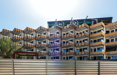 Obraz premium Modern apartment building under renovation with angular balconies, construction equipment on the roof, and a metallic fence in the foreground.