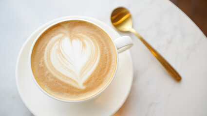 Top view of a creamy latte with elegant heart-shaped foam art in a white cup on marble table, next to a golden spoon glowing softly in natural daylight