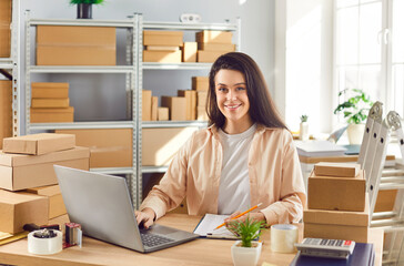Portrait of a woman working in a warehouse, conducting inventory using laptop. She is surrounded by...