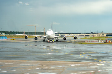 A frontal view of a large Boeing 747 passenger jet on the wet airport tarmac. A pushback tug is attached to the nose gear, conveying aviation logistics under an overcast sky