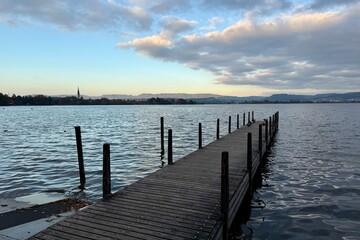 Fototapeta premium Hünenberg am See: A wooden walkway leads out onto Lake Zug, with an autumnal evening atmosphere and clouds in the sky. Symbolic of tranquility, meditation, and peacefulness.