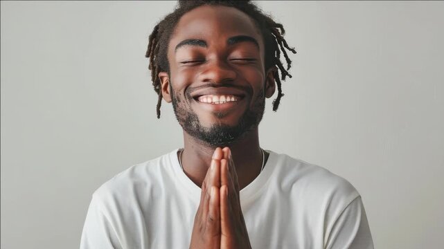 A man with dreadlocks is praying and smiling. He is wearing a white shirt. The shirt is tucked in